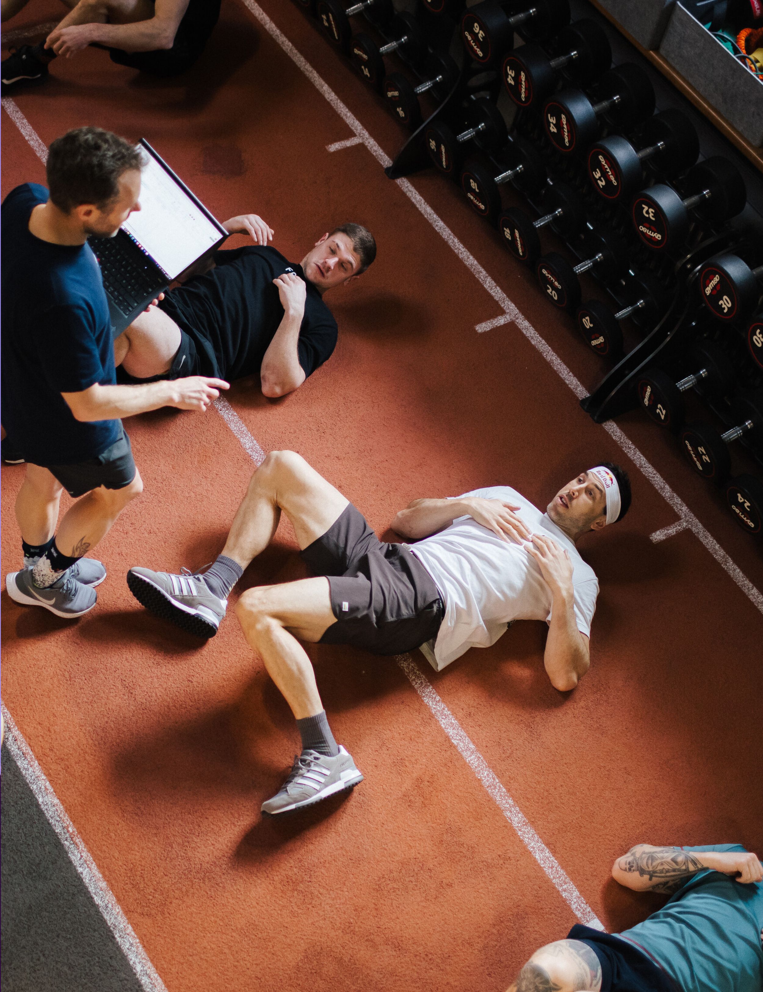 Coach reviewing data on a laptop while two athletes lie on a gym floor recovering beside a rack of dumbbells.