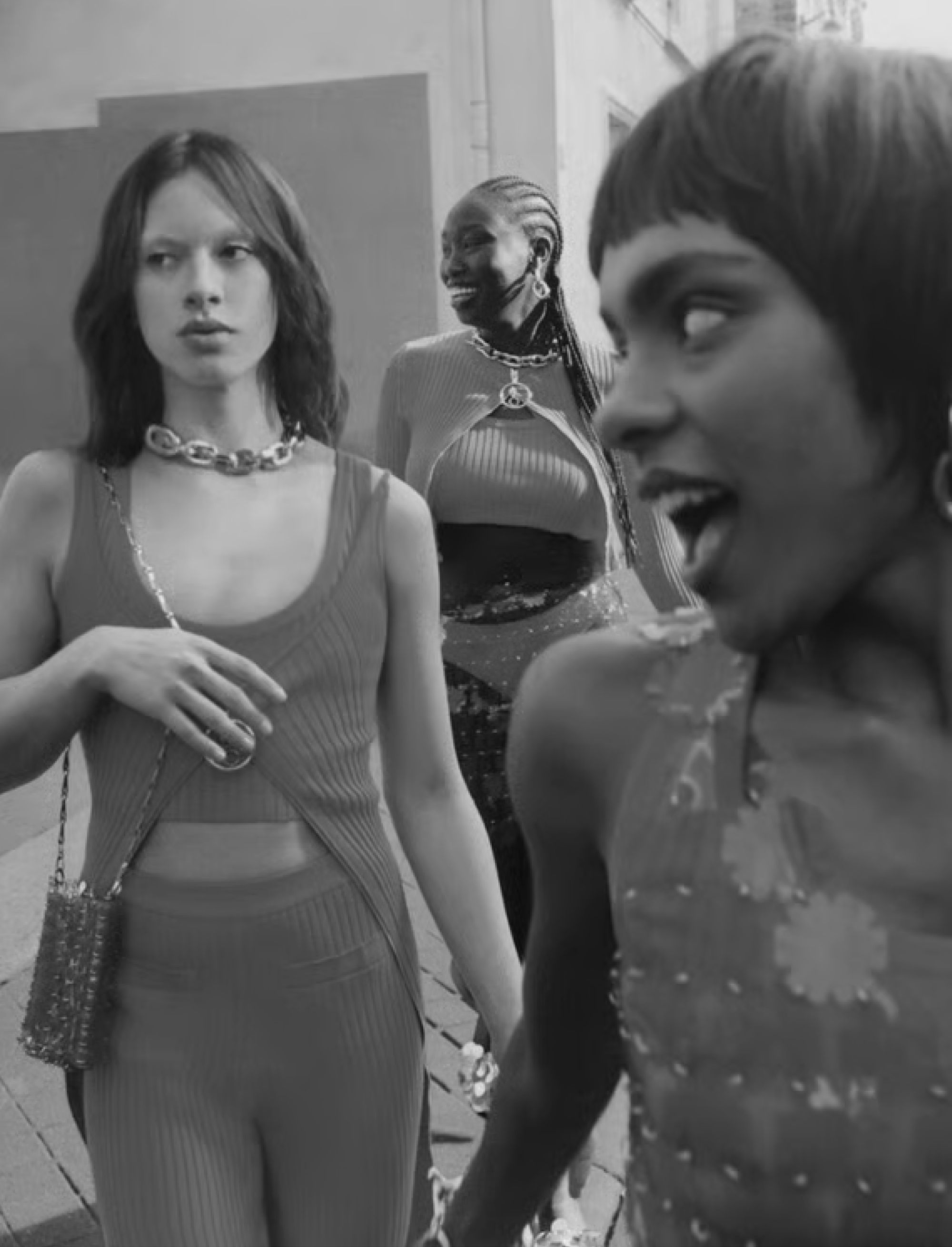 Three fashion models walking together in a black-and-white street scene wearing stylish outfits and jewelry.