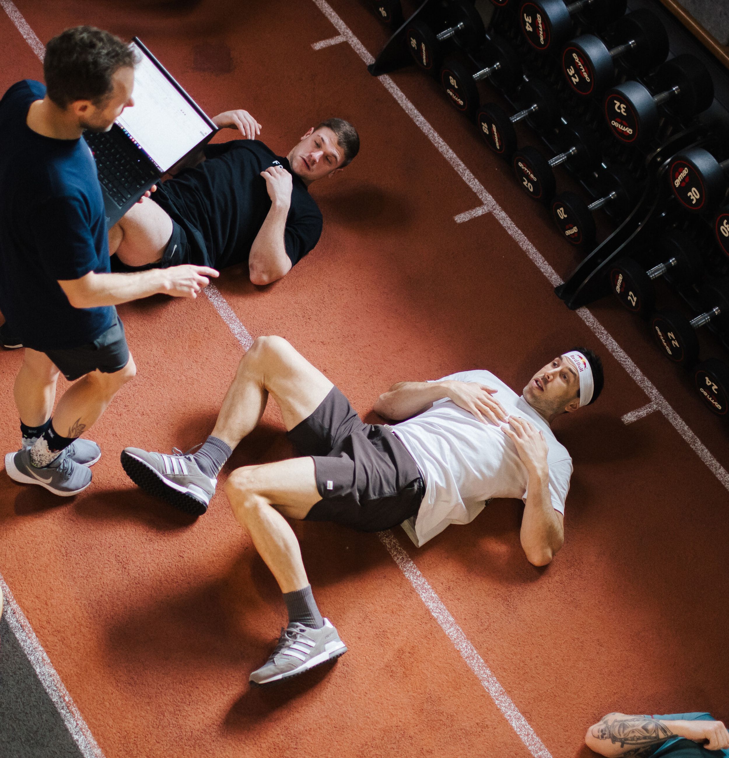 Coach reviewing data on a laptop while two athletes lie on a gym floor recovering beside a rack of dumbbells.