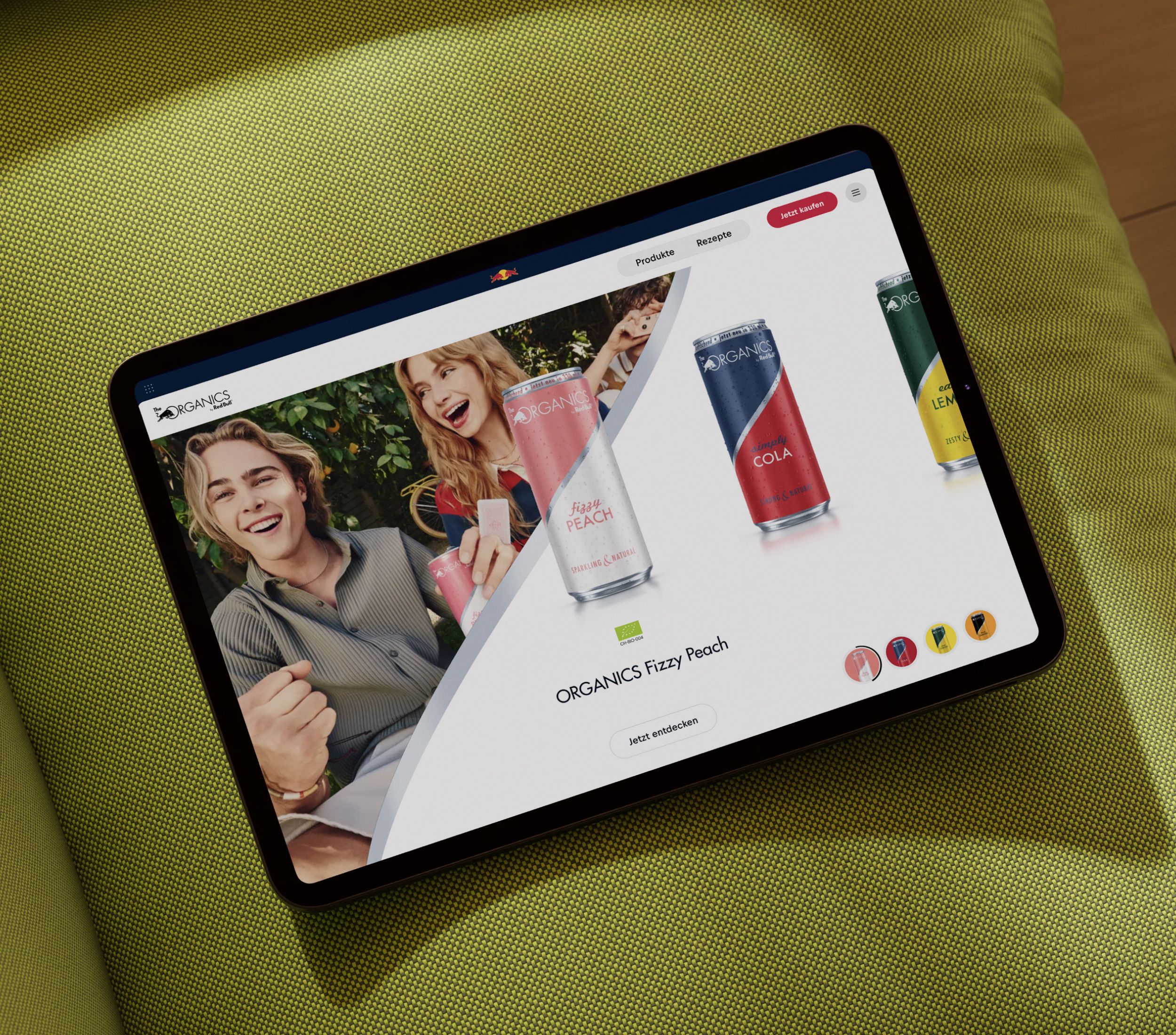 A tablet displaying a website with colorful cans of ORGANICS drinks and people holding beverages, placed on a green textured surface