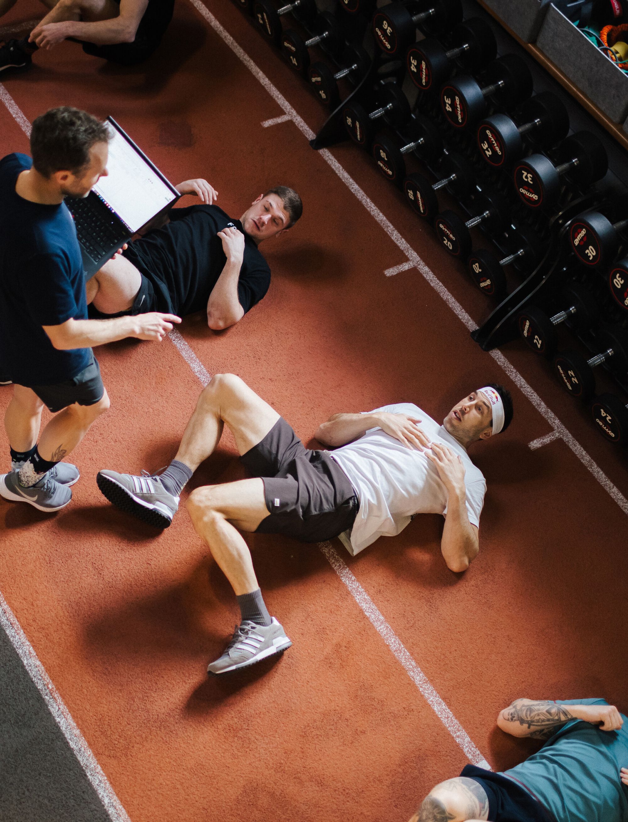 Coach monitoring athletes lying on a gym floor during training beside a rack of dumbbells.