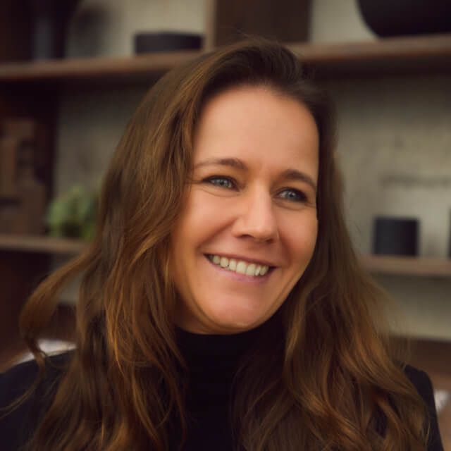 Person with long wavy brown hair, wearing a black turtleneck, standing in front of shelves