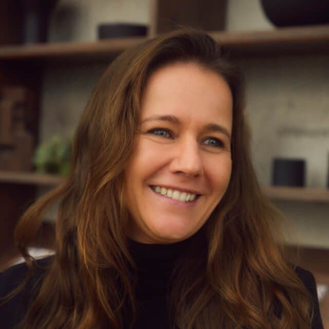 Person with long wavy brown hair, wearing a black turtleneck, standing in front of shelves