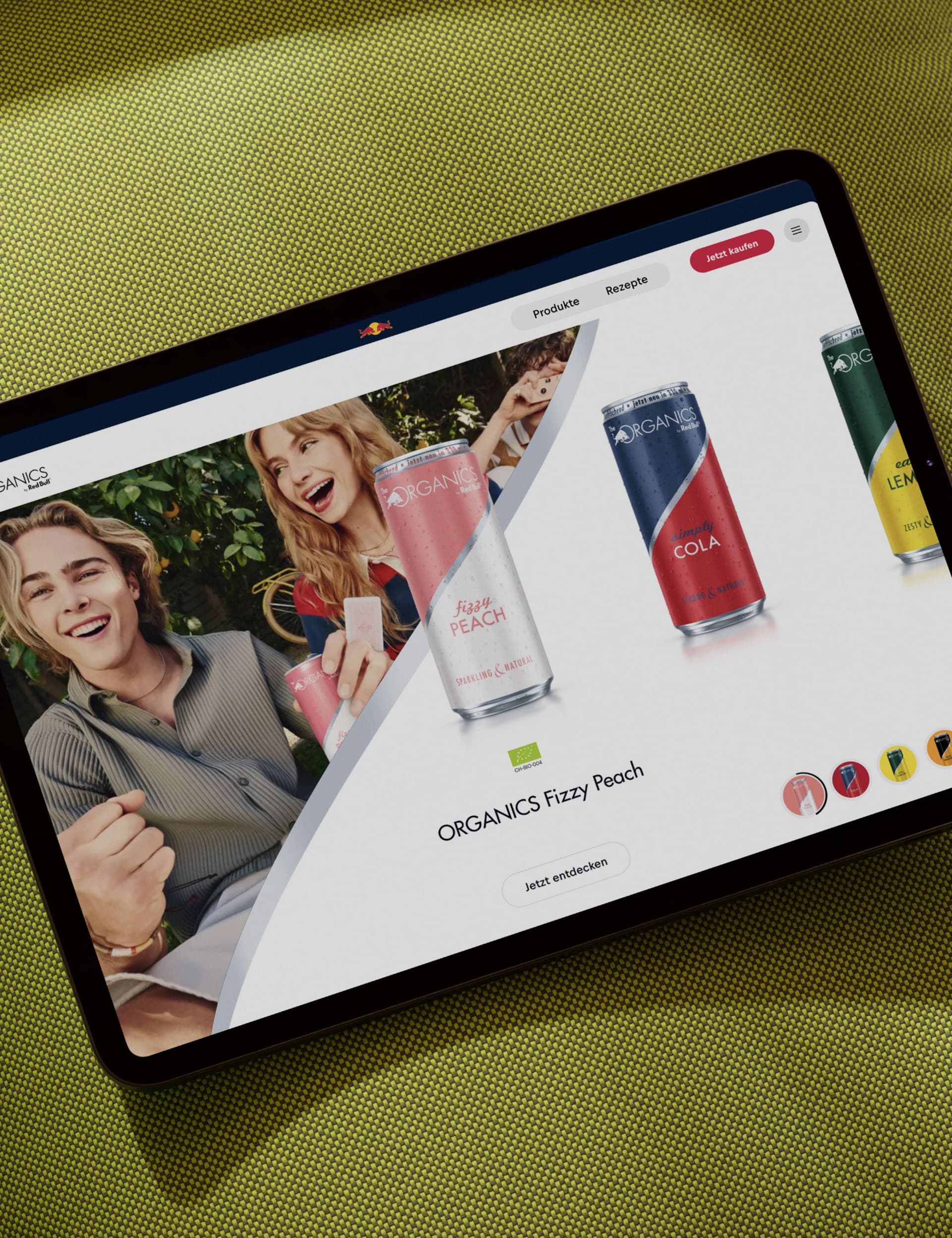 A tablet displaying a website with colorful cans of ORGANICS drinks and people holding beverages, placed on a green textured surface