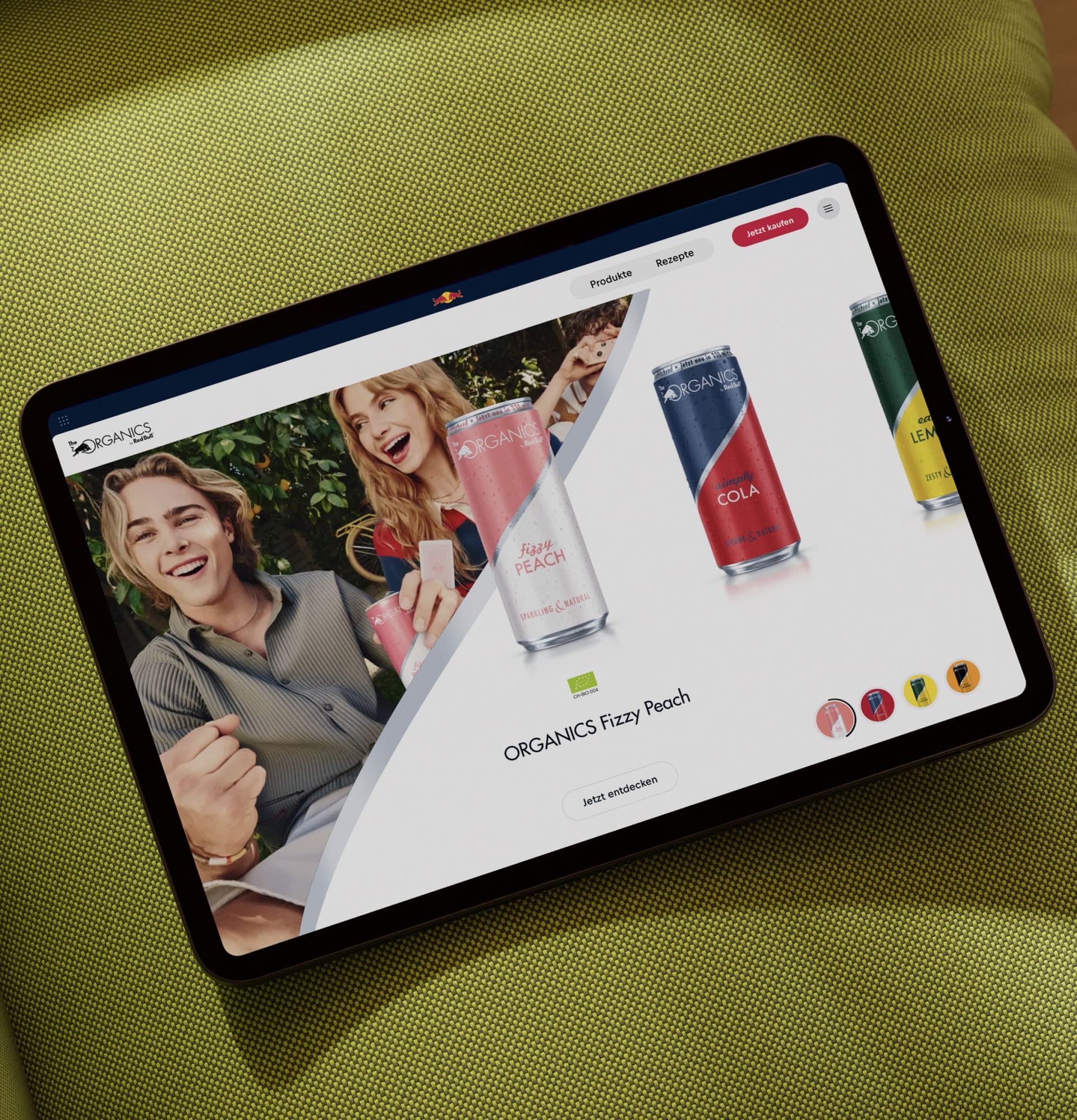 A tablet displaying a website with colorful cans of ORGANICS drinks and people holding beverages, placed on a green textured surface