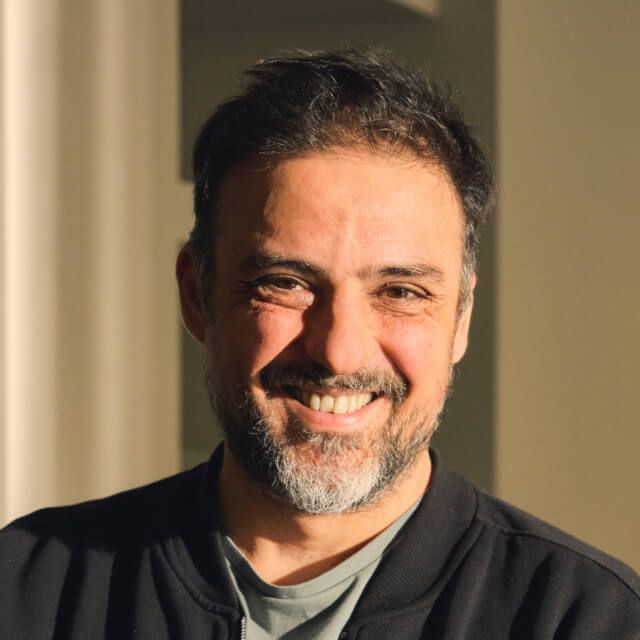 Person with short dark hair and a beard, wearing a black jacket and grey t-shirt, indoors with warm lighting