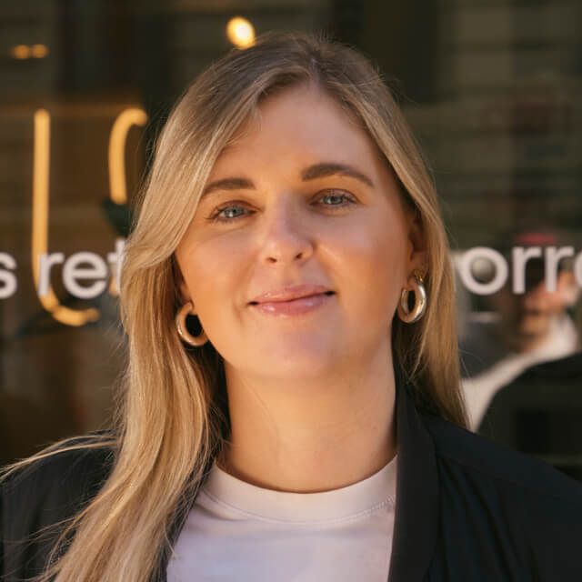 Person with long blonde hair wearing a white top and hoop earrings, standing outdoors in front of a window